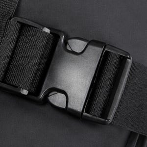 May include: Close-up of a black plastic buckle with a woven nylon strap. The buckle is rectangular with a central release mechanism. The strap is securely fastened within the buckle's frame, and the background is a dark, textured surface.