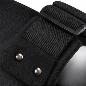 May include: Close-up of a black fabric with a black strap and buckle. Two silver metal rivets are visible on the fabric. The image highlights the texture and construction of the material.