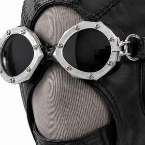 May include: A black leather head covering featuring prominent stitched seams and integrated silver-toned metal goggles. The goggles have dark, circular lenses secured by small screws along the outer rim, with a metallic ring attachment on the side.