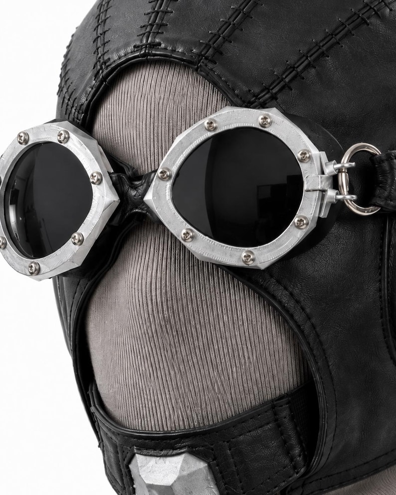 May include: A black leather head covering featuring prominent stitched seams and integrated silver-toned metal goggles. The goggles have dark, circular lenses secured by small screws along the outer rim, with a metallic ring attachment on the side.