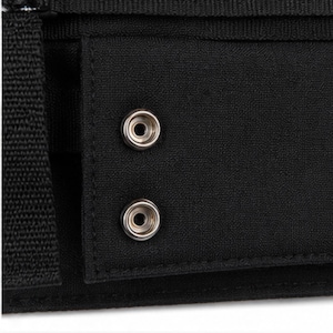 May include: Close-up of a black fabric item with two silver snap closures. The snaps are circular with a raised center and a surrounding ring. The fabric appears to be a durable material, possibly canvas or a similar textile.