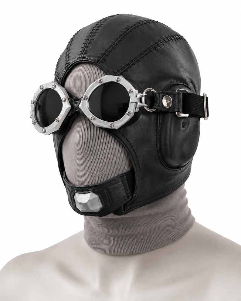 May include: A black leather aviator-style cap featuring stitched paneling and integrated ear flaps. The cap is paired with silver-toned metal goggles that have dark, circular lenses and a chin strap with a central silver-colored geometric accent.