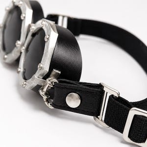 Może przedstawiać: Czarne gogle z dwiema okrągłymi soczewkami w srebrnych metalowych oprawkach z widocznymi nitami. Panele boczne są czarne, połączone z czarnym elastycznym paskiem ze srebrną metalową klamrą oraz czarną sk&oacute;ropodobną wypustką ze srebrnym zatrzaskiem.