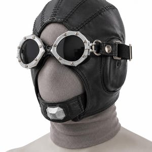 May include: A black leather aviator-style cap featuring stitched paneling and integrated silver-toned metal goggles with dark lenses. The headgear includes a chin strap with a silver-toned accent piece and an adjustable black elastic strap.