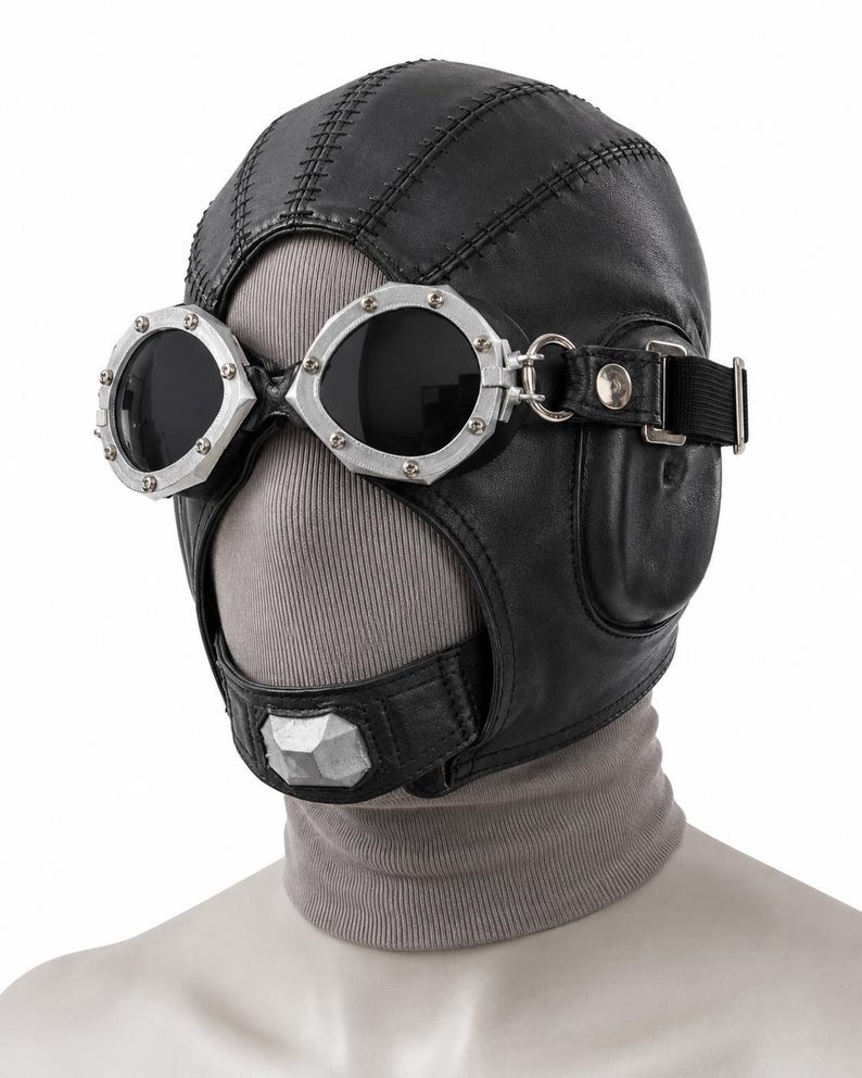 May include: A black leather aviator-style cap featuring stitched paneling and integrated silver-toned metal goggles with dark lenses. The headgear includes a chin strap with a silver-toned accent piece and an adjustable black elastic strap.