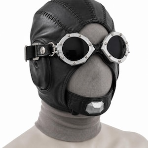 May include: A black leather aviator-style cap featuring prominent stitched seams and integrated black-tinted goggles with silver-toned metal frames. The cap includes a chin strap with a silver-toned metallic accent piece and a grey fabric neck covering.