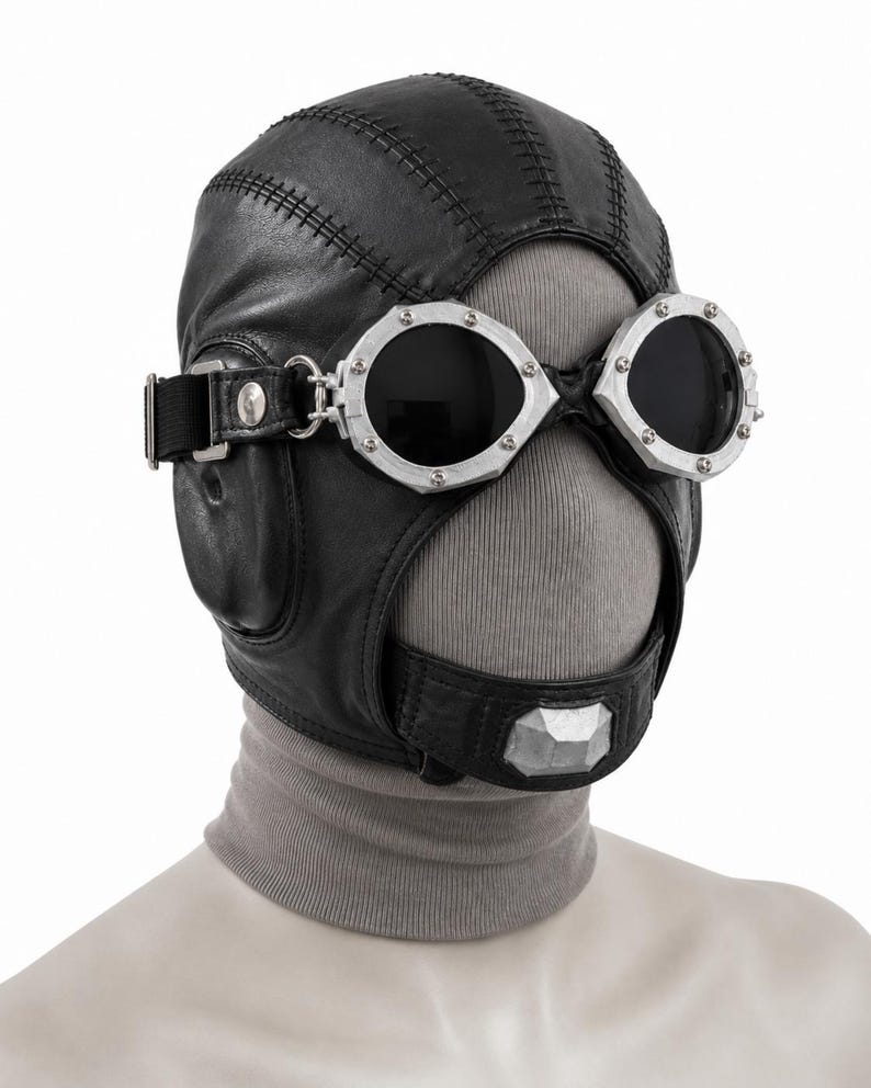 May include: A black leather aviator-style cap featuring prominent stitched seams and integrated black-tinted goggles with silver-toned metal frames. The cap includes a chin strap with a silver-toned metallic accent piece and a grey fabric neck covering.