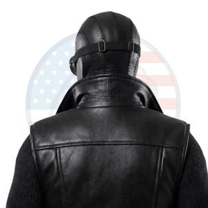 May include: A black leather waistcoat with a high collar worn over a dark ribbed jumper. A black leather head covering with visible stitching and an adjustable elastic strap is positioned on the head, featuring a side-mounted circular lens component.