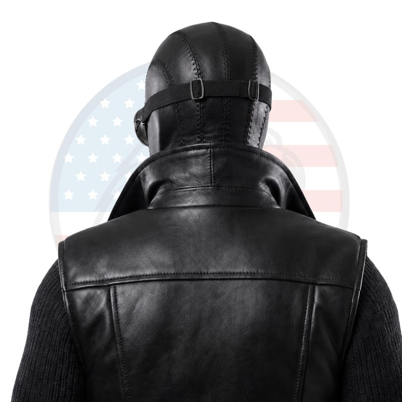 May include: A black leather waistcoat with a high collar worn over a dark ribbed jumper. A black leather head covering with visible stitching and an adjustable elastic strap is positioned on the head, featuring a side-mounted circular lens component.