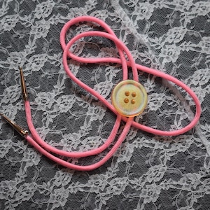 May include: A pink bolo tie with a round, iridescent button featuring four small, gold-colored dots. The tie's cord is a bright pink, and the tips are gold-colored. The background is a white lace pattern.