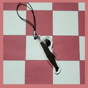 May include: A black and white anime character charm with a black string attached. The charm is of a person in a suit, with their arms raised above their head. The charm is on a checkered background.