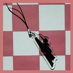 May include: A clear acrylic charm featuring a black silhouette of a person in a hat and coat. The charm is attached to a black cord with a silver-colored metal clasp. The background is a pink and white checkered pattern.