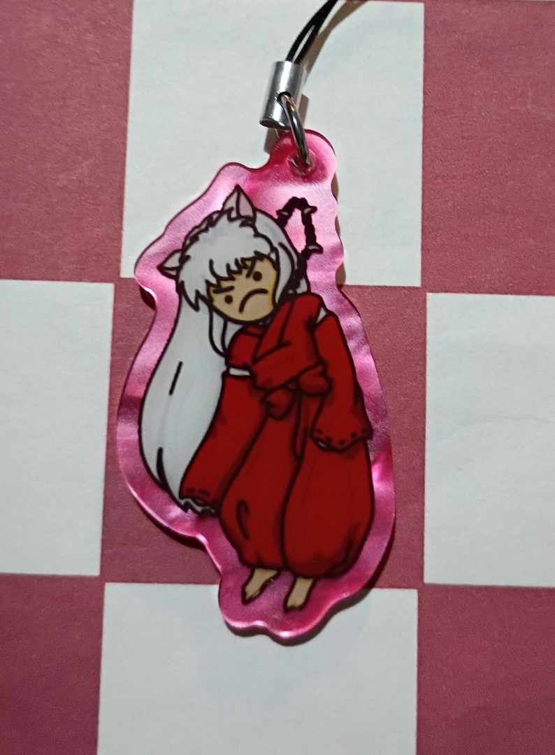 May include: A pink acrylic charm featuring an anime character with white hair and a red outfit. The character has a grumpy expression. The charm is attached to a black lanyard and silver clasp. The background is a pink and white checkered pattern.
