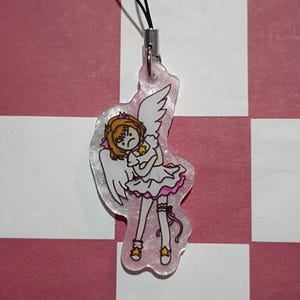 May include: A clear acrylic charm featuring an anime-style character with angel wings, a white dress with pink trim, and star-shaped shoes. The charm is attached to a black cord and silver-colored metal clasp. The background is a pink and white checkered pattern.