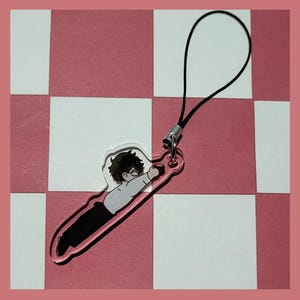 May include: A clear acrylic charm with a cartoon illustration of a person wearing glasses and a white shirt. The charm is attached to a black string.