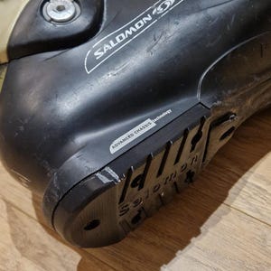 May include: Black and silver Salomon ski boot. The boot features the Salomon logo and "ADVANCED CHASSIS technology" text. The boot is on a wooden surface.