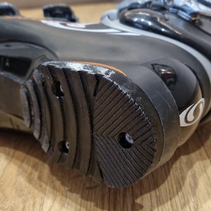 Könnte beinhalten: Nahaufnahme eines schwarzen Skischuhs mit strukturierter, geriffelter Sohle. Der Schuh hat ein glänzendes Finish mit grauen und orangefarbenen Akzenten. Die Sohle weist kreisförmige und winklige Muster für den Halt auf.