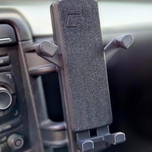 Peut inclure: Support de téléphone de voiture en plastique noir avec une surface texturée. Le support possède deux bras réglables qui peuvent être déplacés pour s'adapter à différentes tailles de téléphone. Le support est conçu pour être fixé à une grille d'aération de voiture.