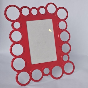 May include: A bright red picture frame with a rectangular opening. The frame is decorated with a series of connected circles of varying sizes around the perimeter. The frame is made of a glossy material.