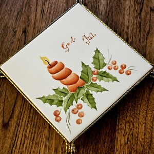Può includere: Piastrella quadrata in ceramica con cornice in metallo dorato. La piastrella presenta un disegno dipinto a mano di una candela, foglie di agrifoglio e bacche. Le parole "God Jul" sono scritte in arancione. Una decorazione festiva natalizia.