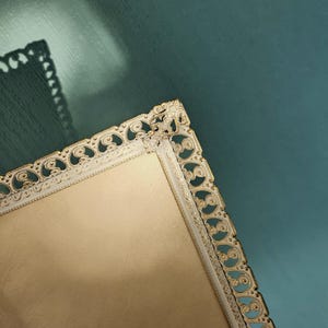 May include: A rectangular, cream-colored tray with a decorative white and gold filigree border. The tray has a soft, textured surface and is set against a teal background. The ornate design adds a touch of elegance.