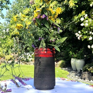 May include: A handmade ceramic vase with a black and red design, filled with a vibrant arrangement of yellow and purple wildflowers. The vase is cylindrical and sits on a white surface, with a lush green garden in the background.