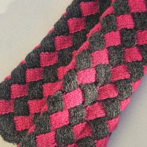 May include: Close-up of a knitted scarf with a checkerboard pattern. The scarf features alternating squares of dark grey and bright pink. The texture of the knit is visible, creating a cosy and handmade appearance.