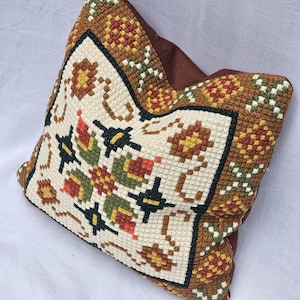 Può includere: Cuscino quadrato decorativo con un dettagliato motivo a punto croce. Il cuscino presenta un centro color crema con un motivo floreale rosso, verde, giallo e nero. Il bordo è marrone con ulteriori motivi floreali e geometrici. Il retro del cuscino è marrone tinta unita.
