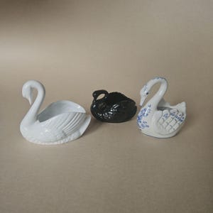 Peut inclure: Trois figurines de cygnes en céramique, en blanc, noir et blanc avec des motifs floraux bleus. Les cygnes sont conçus comme de petits récipients. Le cygne blanc de gauche a des détails de plumes.