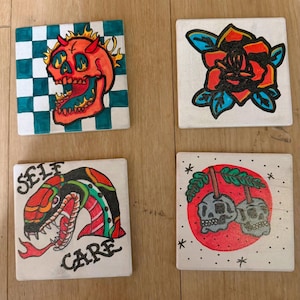 May include: Four square ceramic coasters with tattoo-style designs. One features a red skull with flames on a checkered background. Another shows a red rose with blue leaves. A third has a snake with the words "SELF CARE". The last has two skulls.