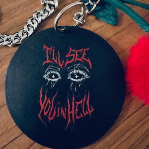 May include: Black round keychain with the text "I'll See You in Hell" in red gothic lettering. The keychain features a drawing of eyes with tears. A silver chain and a red fluffy pom-pom are attached.