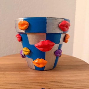 May include: A hand-painted ceramic flower pot with a blue and silver checkered pattern. The pot is adorned with colorful, three-dimensional lip designs in shades of red, orange, yellow, and purple. The pot sits on a wooden surface.