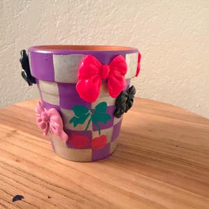 Puede incluir: Una maceta decorativa con un patrón a cuadros en morado y blanco. La maceta está adornada con lazos rosas, negros y rosas. Tréboles verdes y cerezas rojas están pintados en la maceta.