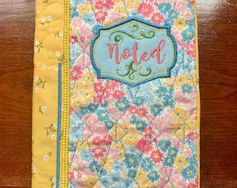 May include: A quilted book cover with a floral pattern in shades of pink, blue, and yellow. The cover has a blue embroidered patch with the word "Noted" in a decorative font.