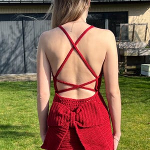 Op de afbeelding: Een rode gehaakte jurk met een gekruiste rug en een grote strik in de taille. De jurk is gemaakt van getextureerd garen en heeft een getailleerd silhouet. De jurk is knielang en geschikt voor een speciale gelegenheid.