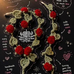 Op de afbeelding: Een gehaakte kabelbeschermer met saliegroene bladeren en felrode rozen. De tekst bevat "handmade with love," "little details, big happiness," "soft, cozy & made with so much love," "cute little roses," "every stitch made with care," en "Stitch & Glam By Kiana."