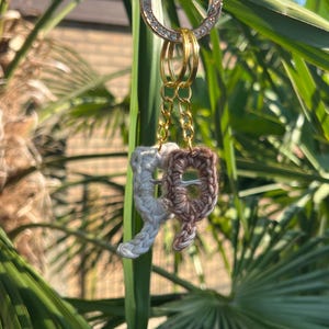 Op de afbeelding: Een goudkleurige sleutelhanger met een cirkelvormige, met strass bezette bedel en twee gehaakte letterbedels. Eén letter is wit en de andere is bruin. De sleutelhanger hangt voor groen gebladerte.