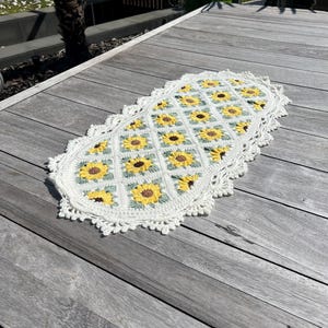Op de afbeelding: Een wit gehaakt ovaal vloerkleed met een zonnebloemmotief. Het vloerkleed ligt op een houten oppervlak.