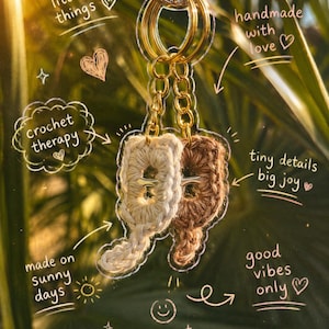 Op de afbeelding: Een sleutelhanger met twee kleine, gehaakte katvormige bedels in crème en bruin garen, bevestigd aan een goudkleurige metalen ring. De afbeelding bevat handgeschreven tekst: soft little things, handmade with love, crochet therapy, tiny details big joy, made on sunny days, good vibes only en you got this.