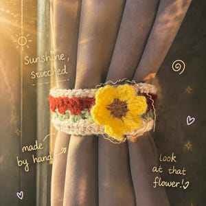 Op de afbeelding: Een gehaakte gordijnophouder met een horizontale band van crème, rood en lichtgroen garen, versierd met een opvallende gele bloem met een bruin hart. De tekst "Sunshine, Stitched", "made by hand" en "look at that flower!" is zichtbaar.