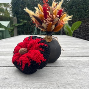 Op de afbeelding: Een zwarte vaas houdt een boeket gedroogde bloemen in tinten rood, oranje en geel vast. Een gehaakte pompoen in rood en zwart staat voor de vaas. De pompoen heeft een bruine steel.