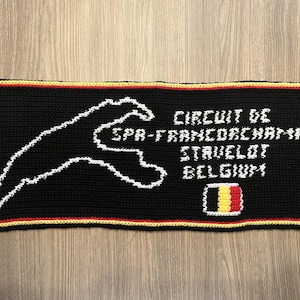 Op de afbeelding: Zwart gehaakte rechthoek met witte omtrek van een racecircuit en de tekst "CIRCUIT DE SPA-FRANCORCHAMPS, STAVELOT, BELGIË". Een kleine Belgische vlag staat in de hoek. De rand is rood, geel en zwart.