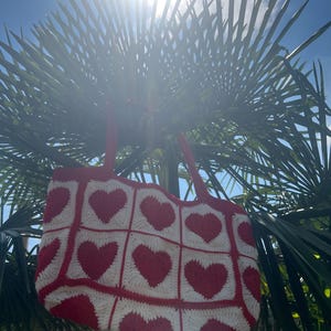 Op de afbeelding: Een rood-witte gehaakte tote bag met een hartpatroon. De tas heeft een rechthoekige vorm en rode handvatten. Hij hangt aan een palmboom, met de heldere zon op de achtergrond.