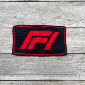 Op de afbeelding: Een zwart en rood gehaakte patch met het F1-logo. Het rode logo heeft een rode rand op een zwarte achtergrond. De rechthoekige patch lijkt handgemaakt.