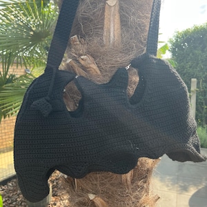 Op de afbeelding: Een zwarte gehaakte tas in de vorm van een vleermuis, met een lange schouderriem. De tas heeft uitgesneden details en een getextureerde, handgemaakte uitstraling. De tas hangt aan een boomstam.