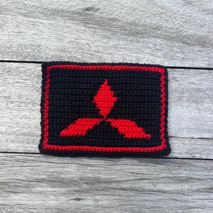 Op de afbeelding: Een zwart gehaakt embleem met een rode rand en een rood drievoudig diamantlogo. De rechthoekige patch lijkt handgemaakt. Het logo is een gestileerd klaverbladontwerp.