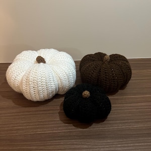 Op de afbeelding: Drie gehaakte pompoenen in wit, bruin en zwart, elk met een bruine steel. De pompoenen zijn gerangschikt op een bruin houten oppervlak, met een herfst oogst thema. De pompoenen variëren in grootte.