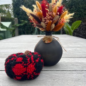 Op de afbeelding: Een zwarte keramische vaas met een arrangement van gedroogde bloemen en een gehaakte pompoen. Het arrangement bevat rode, oranje en bruine gedroogde bloemen. De pompoen is zwart en rood, met een bruine steel.