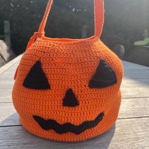 Op de afbeelding: Een oranje gehaakte pompoenvormige tas met een handvat. De tas heeft zwarte driehoekige ogen en een gebogen mond, waardoor een pompoengezicht ontstaat. Perfect voor Halloween, voor snoep of als decoratie.