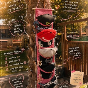 Op de afbeelding: Een roze gehaakte wandorganizer met meerdere vakken voor diverse baseballpetten en haarklemmen. De organizer hangt aan een houten stok en is voorzien van handgeschreven teksten zoals little things made by hand en hat collection.